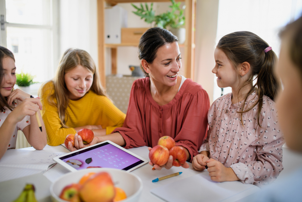 A young homeschooling mom and two daughters are working on school work with fruit at the kitchen table.